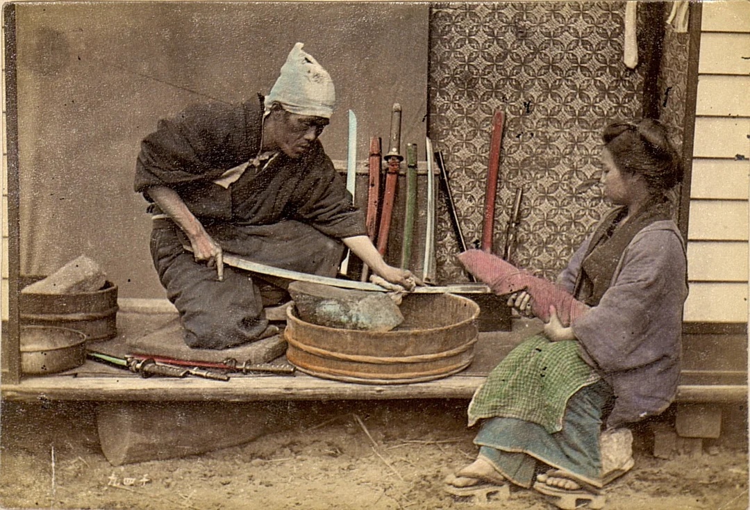 A man sharpens a samurai sword over a wooden basin while a woman sits nearby holding a bundle. Several swords rest against a patterned wall behind them, evoking a historical Japanese setting.
