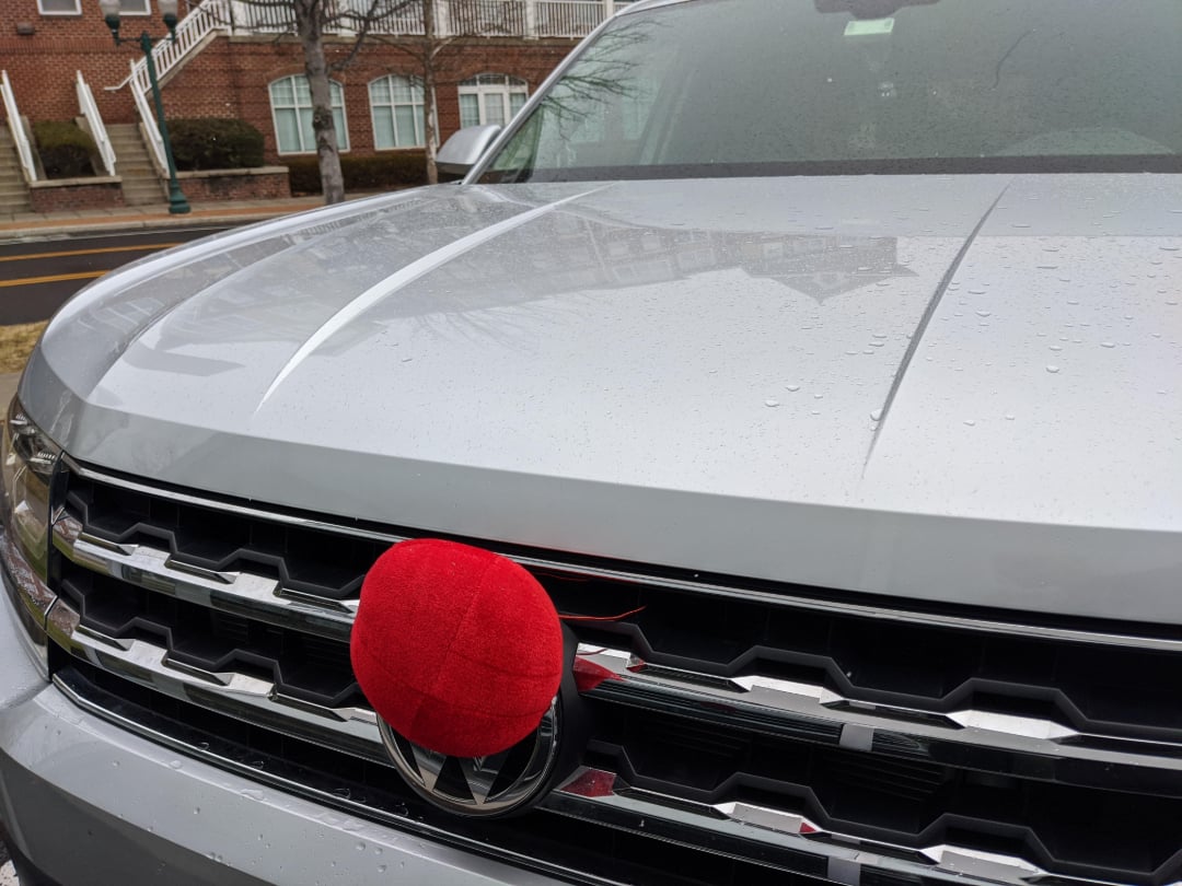A silver car with raindrops on the hood has a large red clown nose attached to the front grille. A brick building and bare trees are reflected on the car’s surface in the background.