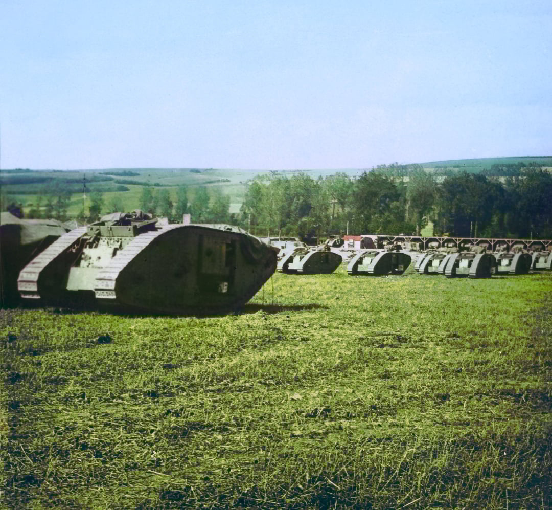 A row of World War I tanks is lined up on a grassy field, with rolling hills and trees in the background under a clear sky.