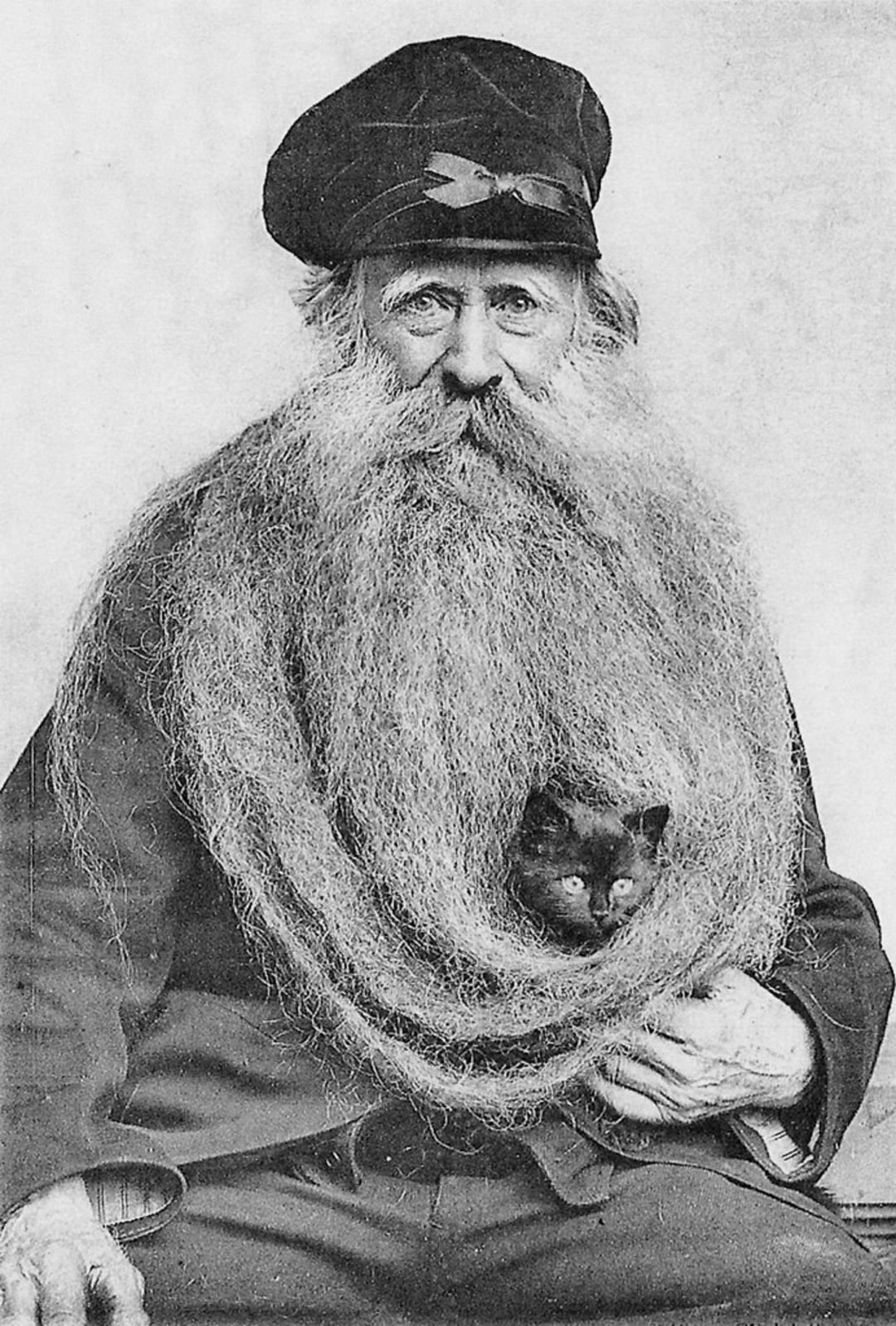 An older man with a very long, bushy beard wears a cap and suit. A small cat is nestled comfortably within the folds of his beard, peeking out toward the camera. The image is in black and white.