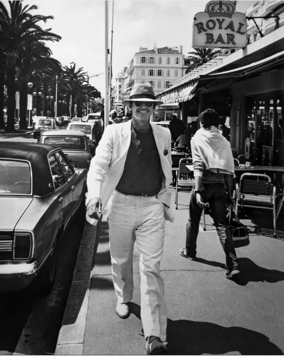 A man in a light-colored suit, hat, and sunglasses walks down a lively city street lined with palm trees and parked cars, passing a cafe called “Royal Bar.” The photo is black and white, evoking a vintage atmosphere.