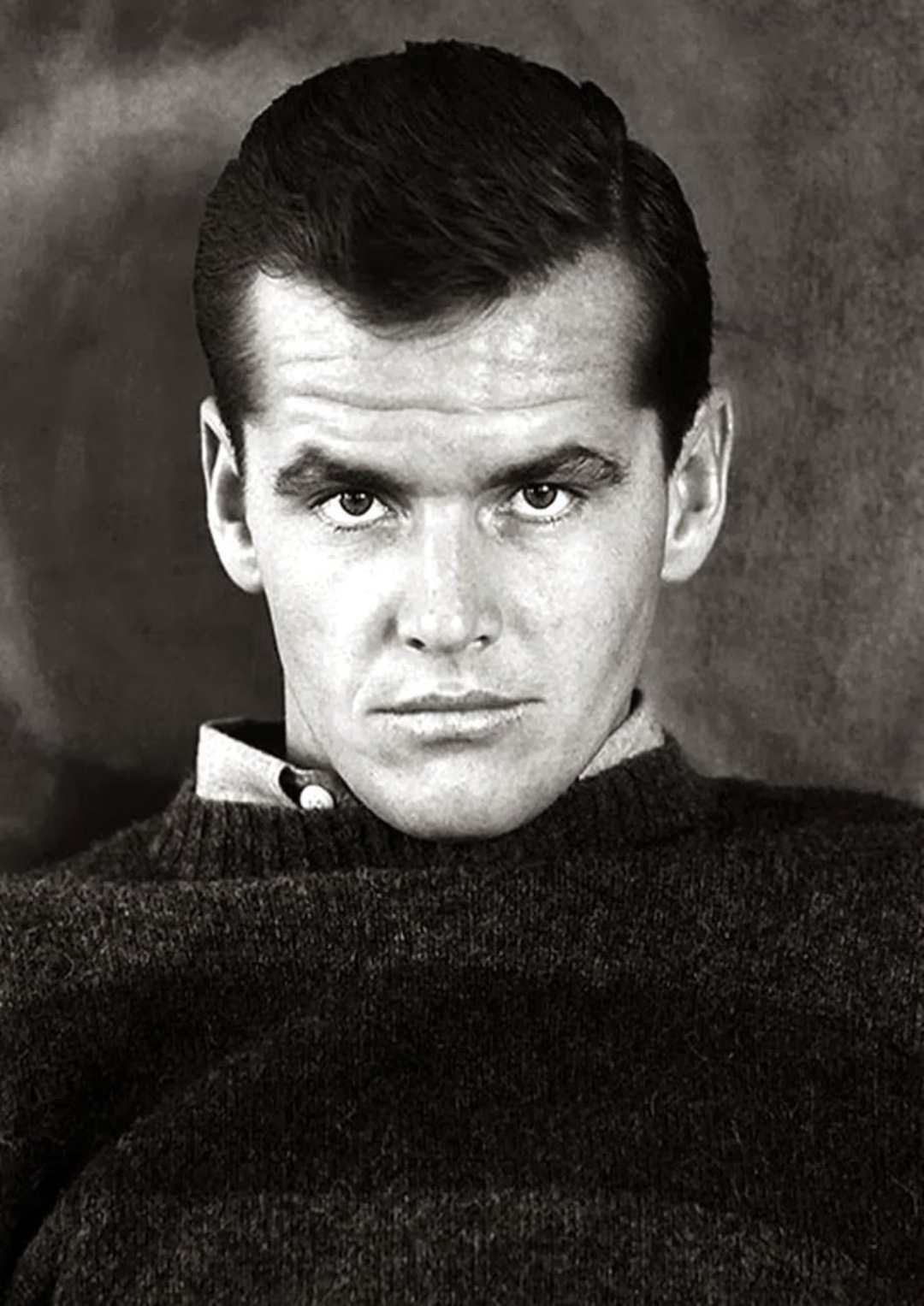A young man with neatly combed dark hair stares directly at the camera, wearing a collared shirt under a dark sweater against a plain, dark background.