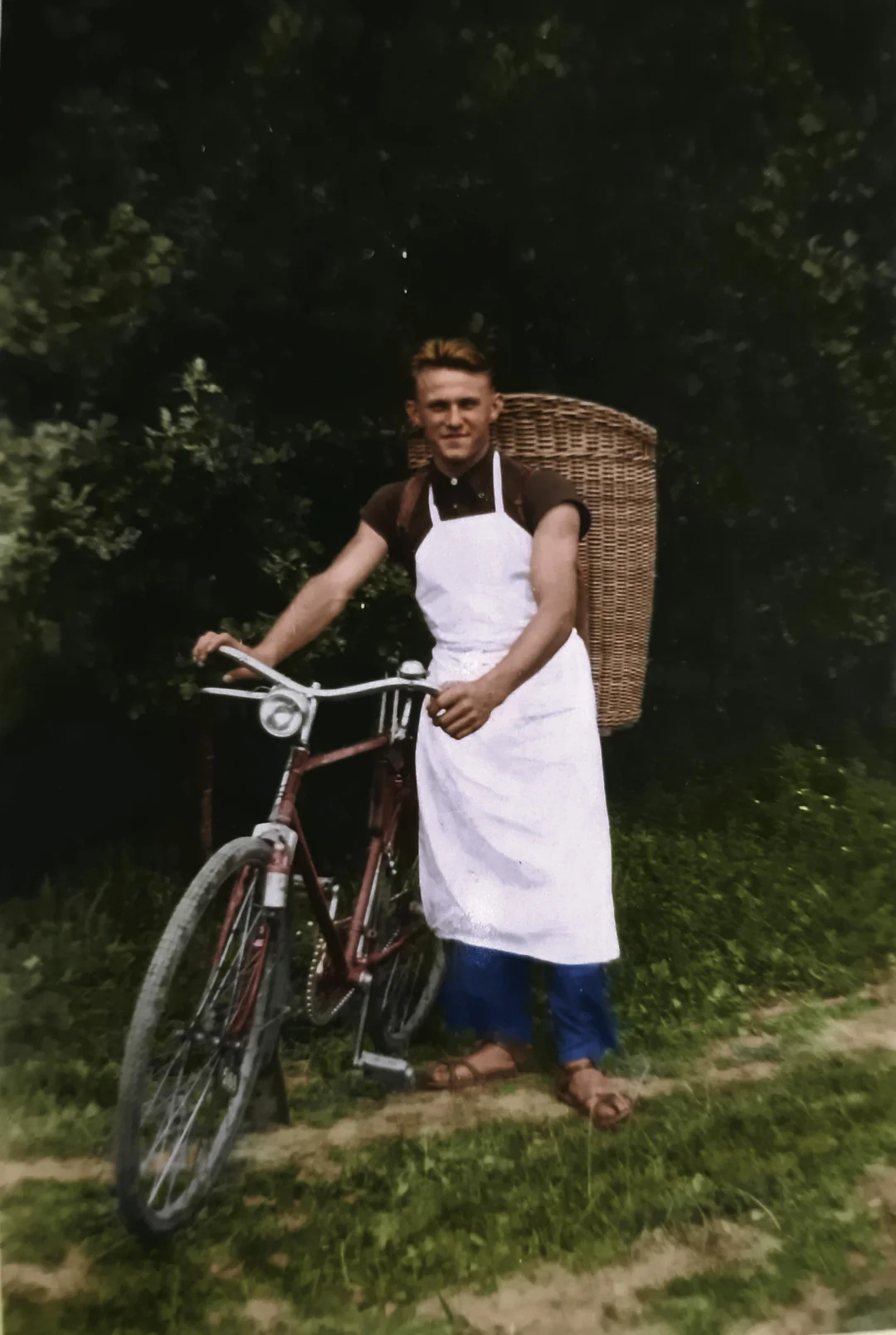 A man wearing a white apron and blue pants stands outdoors beside a bicycle, holding its handlebars. He has a large wicker basket strapped to his back, with green trees and grass in the background.