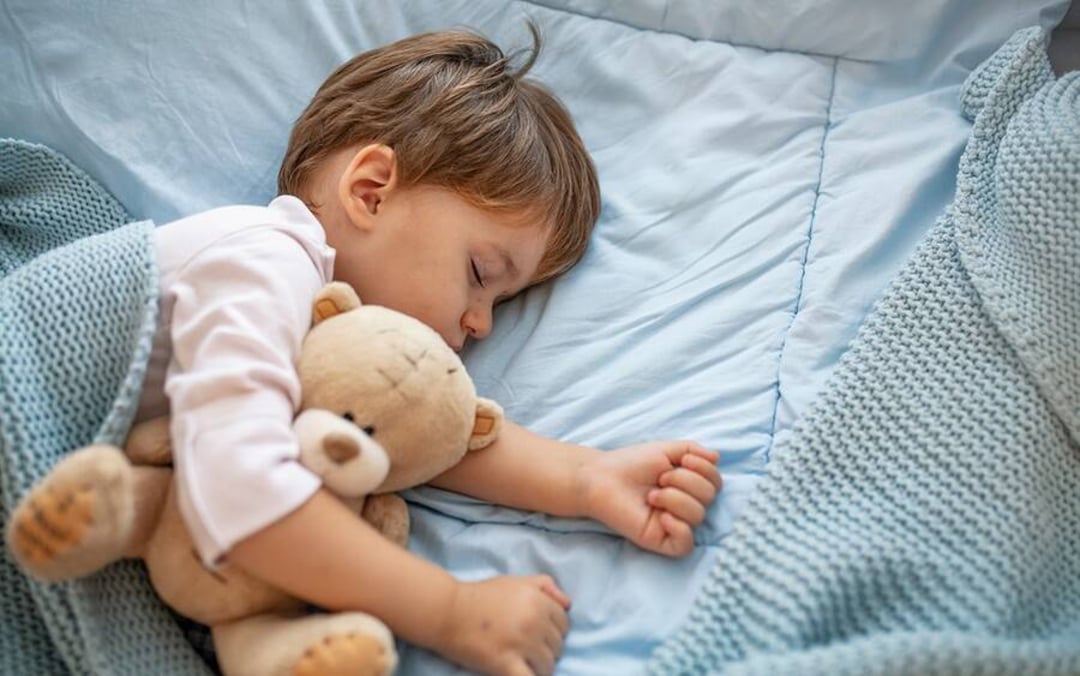 A young child sleeps peacefully in bed, cuddling a brown teddy bear, covered with a light blue blanket. The bedding and blanket are also light blue, creating a calm, cozy atmosphere.