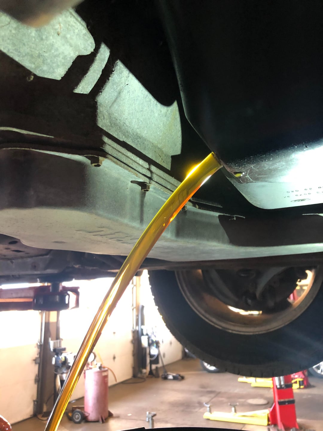 Golden engine oil pours from the underside of a vehicle during an oil change in a garage, with a tire and automotive equipment visible in the background.