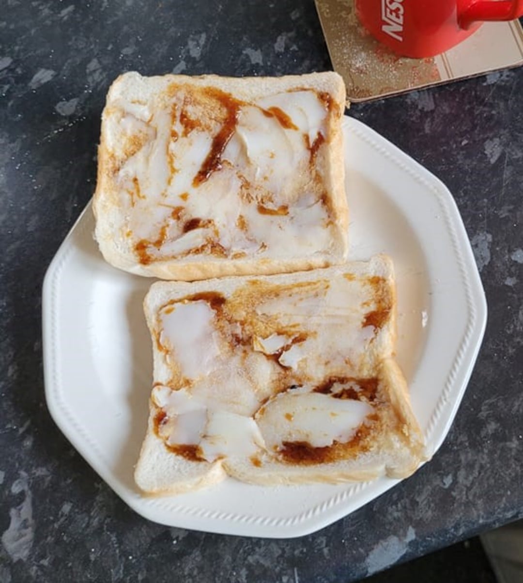 Two slices of white bread on a white plate, each spread with a layer of white fat and streaked with brown sauce. A red mug sits in the upper right corner on the grey countertop.
