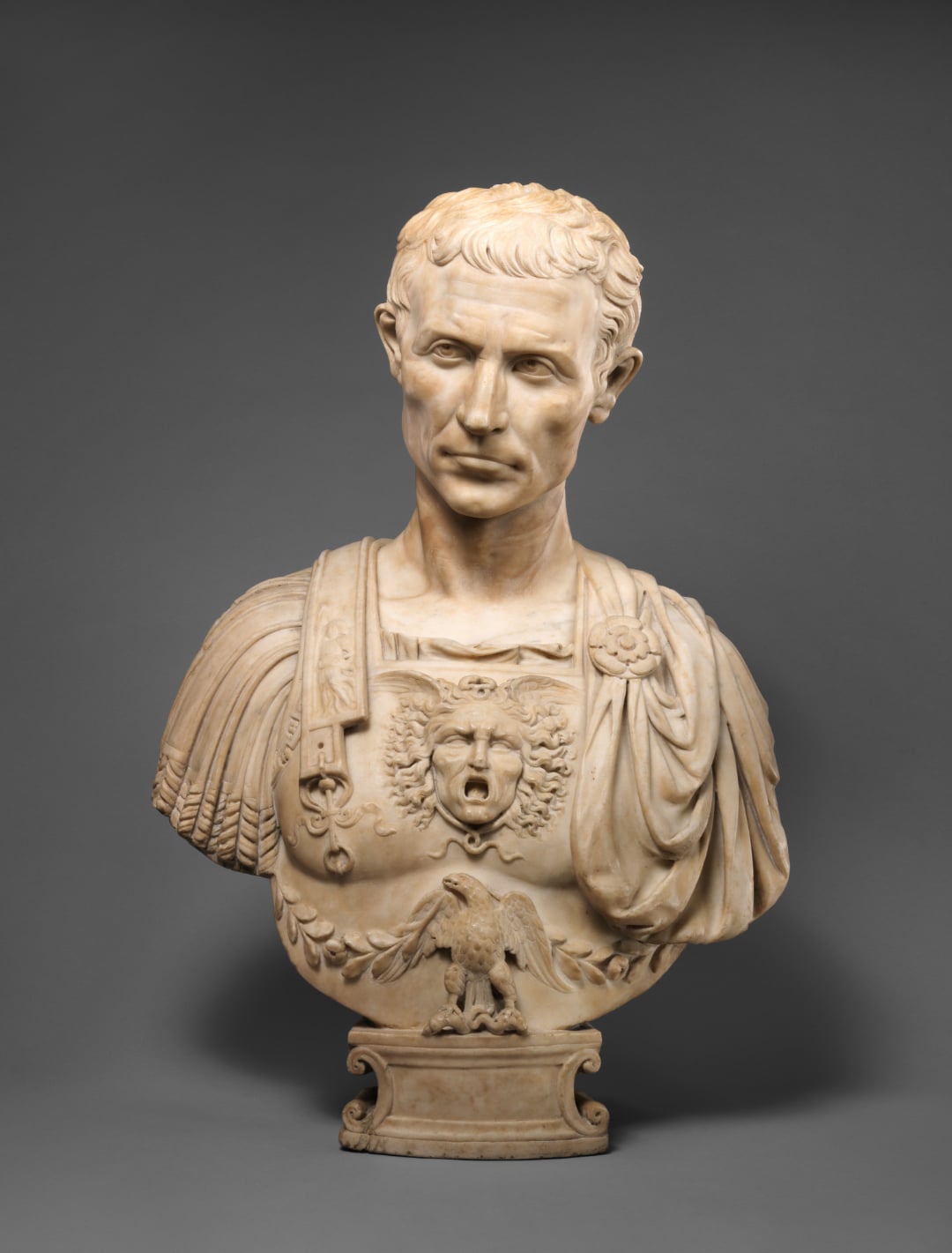 Marble bust of a man with short hair and a serious expression, wearing detailed Roman armor adorned with a relief of Medusa’s head and an eagle at the base, set against a plain gray background.