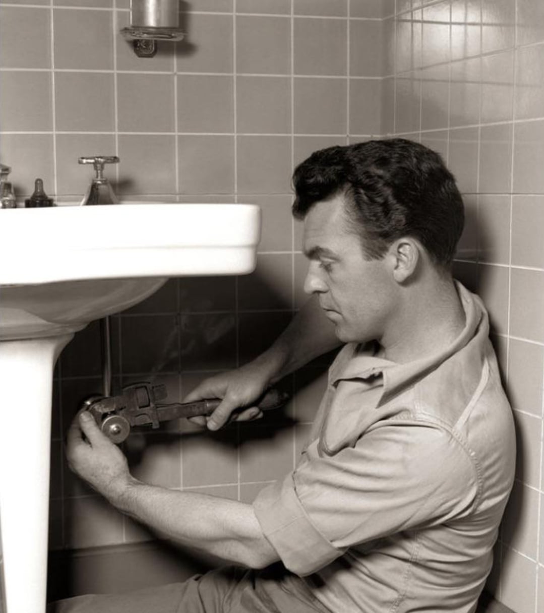 A man in work clothes uses a large wrench to fix pipes beneath a bathroom sink, surrounded by tiled walls.