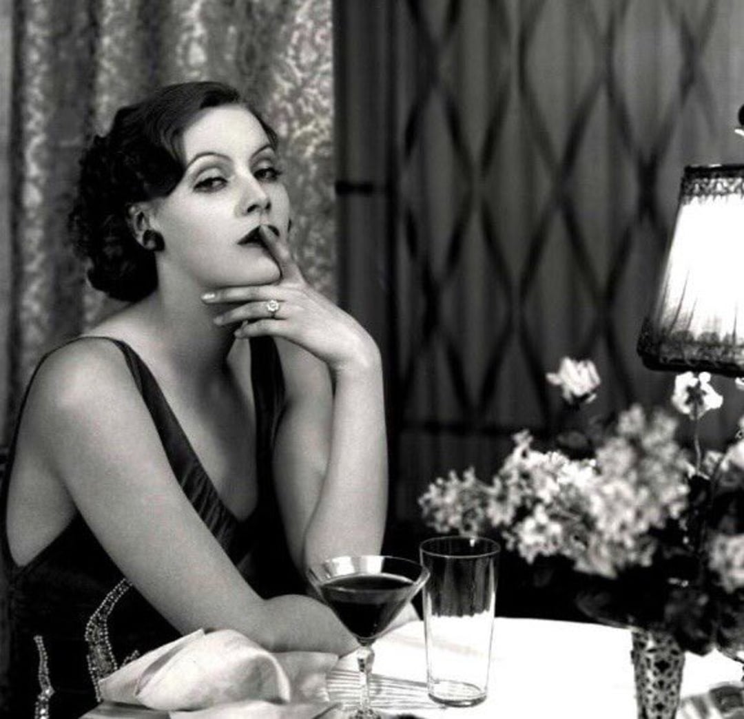 A woman in a glamorous evening dress sits at a table, holding a cigarette to her lips. A cocktail, glass, and a floral arrangement are on the table. Soft light from a lamp creates a vintage, elegant ambiance.