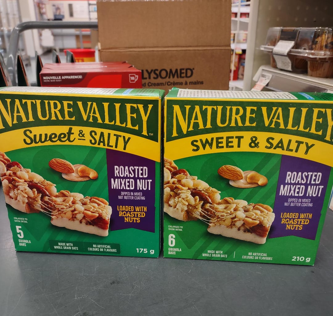 Two boxes of Nature Valley Sweet & Salty Roasted Mixed Nut granola bars are on a counter; one box contains 5 bars (175g) and the other contains 6 bars (210g). Store shelves and products are visible in the background.