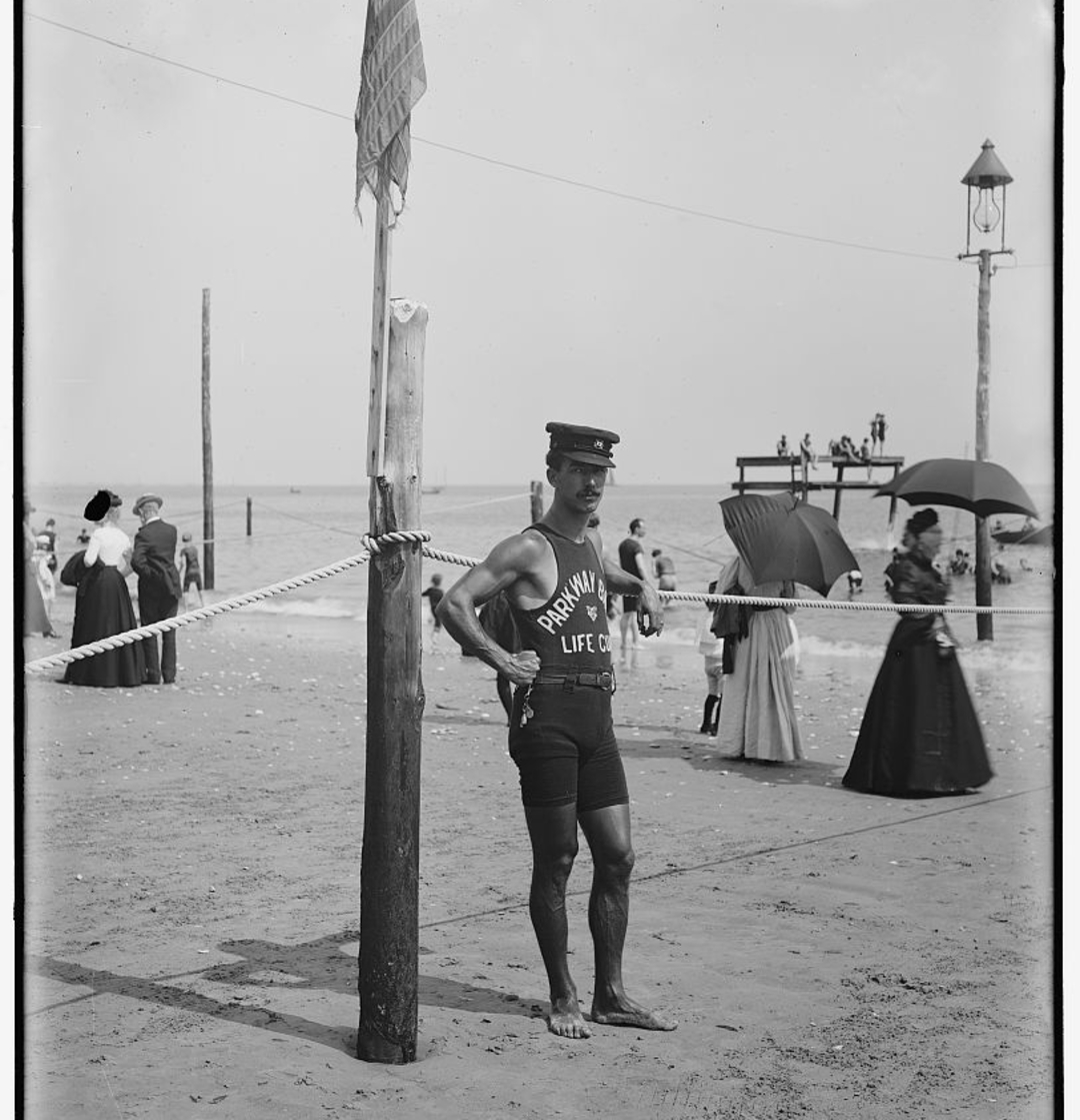 A vintage photo of a lifeguard in a dark uniform and cap stands on a sandy beach near a flagpole and rope, while people in early 1900s attire walk and gather by the shoreline in the background.