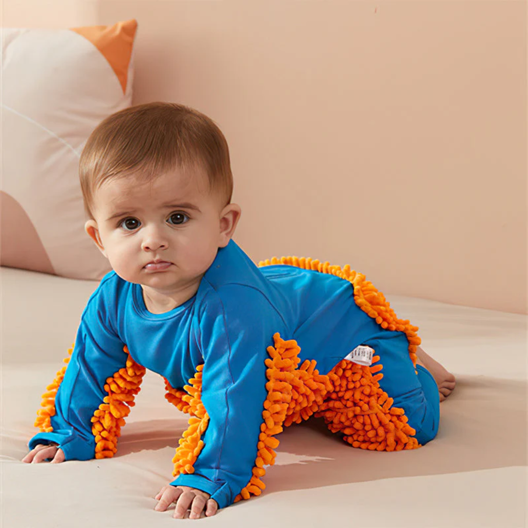 A baby in a blue outfit with bright orange textured fringe crawls on a bed with light-coloured bedding and pillows in the background.