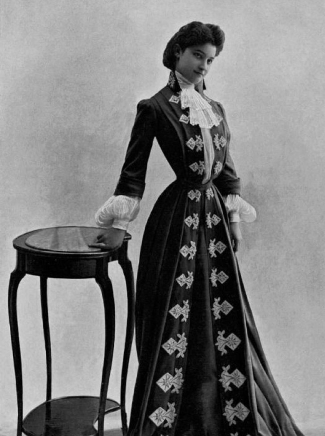 A woman in a long, dark, ornate dress with embroidered patterns stands beside a small round table, posing for a vintage black-and-white portrait.