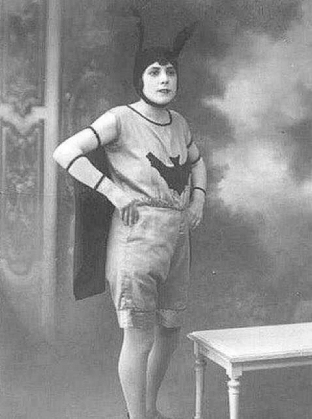 A person in an old-fashioned, homemade bat-themed costume stands with hands on hips. The costume features a bat symbol on the chest, a cape, and headpiece with two tall points. The photo is black and white and vintage in style.