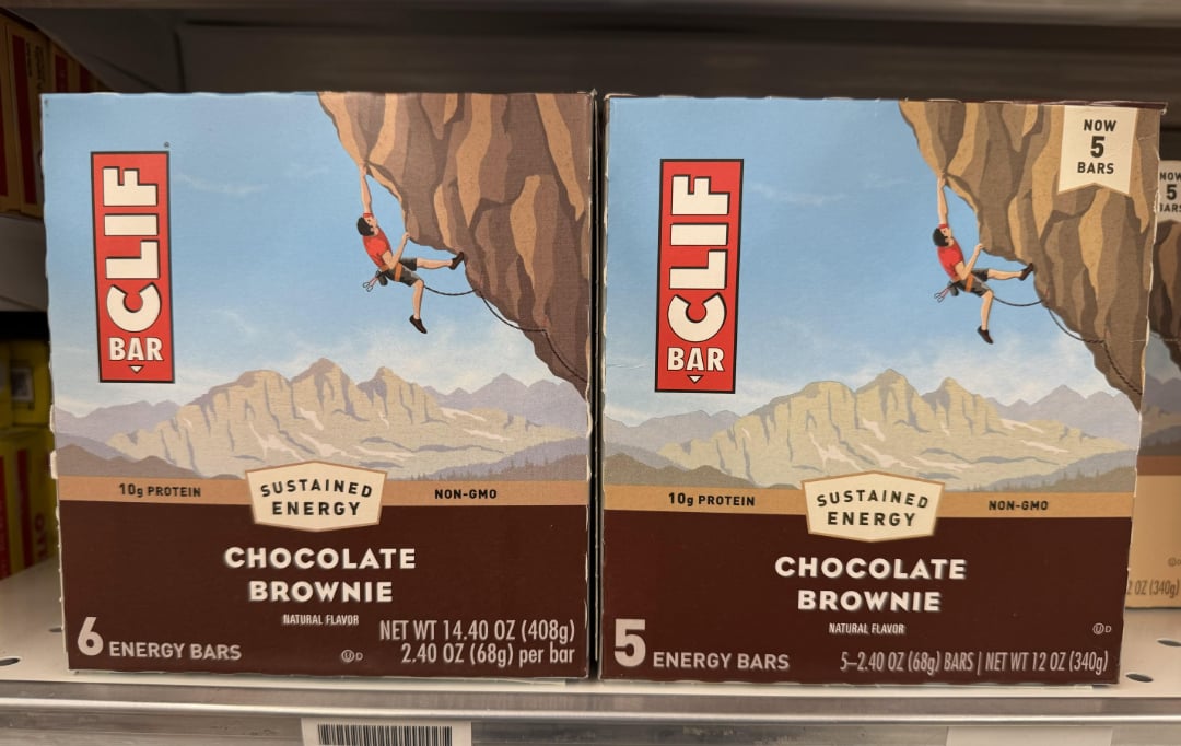Two boxes of Clif Bar Chocolate Brownie energy bars are on a shelf. Each box features an illustration of a climber scaling a rocky cliff with mountains in the background. One box contains 6 bars, the other contains 5.