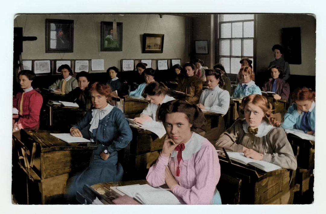 A group of young women sit at wooden desks in a classroom, writing or looking toward the camera. The classroom has framed pictures on the walls, large windows, and a teacher standing at the back.