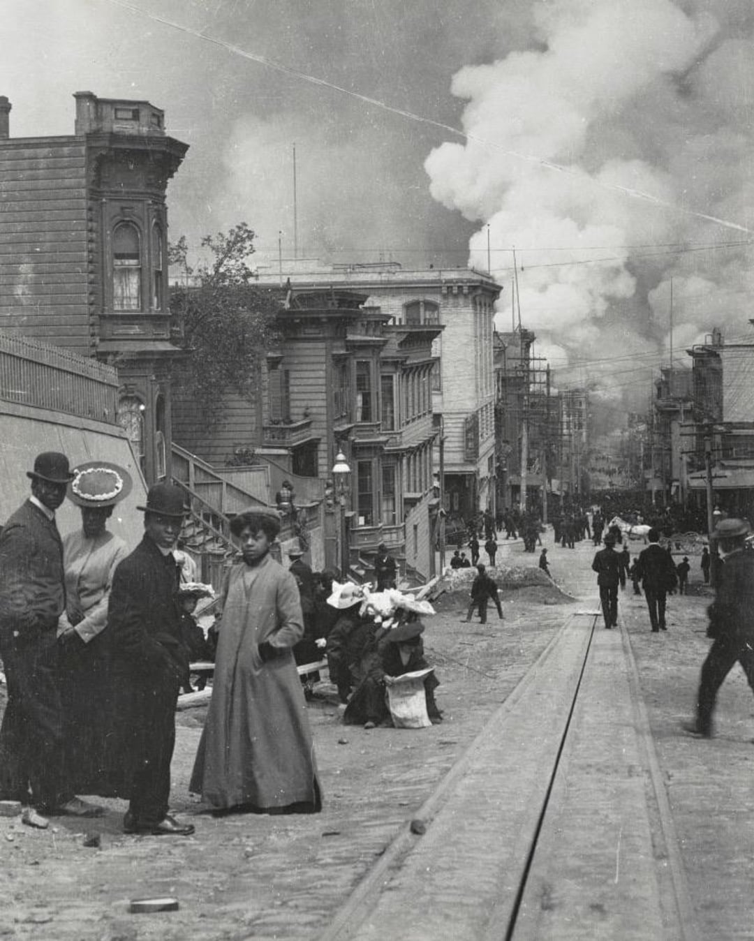 Black-and-white photo of people in early 1900s attire standing and walking on a city street, with smoke rising from buildings in the background. The street is lined with wooden houses and tram tracks.
