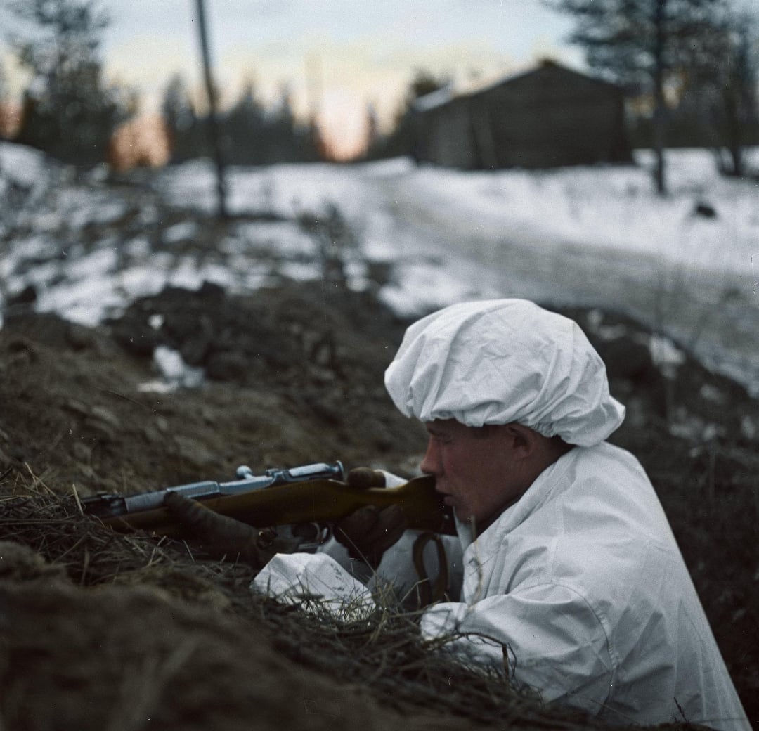 A person dressed in white winter camouflage crouches in a snowy trench, aiming a rifle. Snow-covered ground and a wooden building are visible in the background, suggesting a cold, outdoor setting.