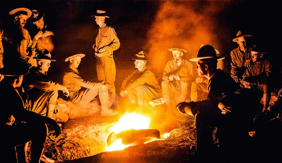 A group of men in uniform sit and stand around a glowing campfire at night, their faces illuminated by the firelight as they talk and relax together, wearing wide-brimmed hats.