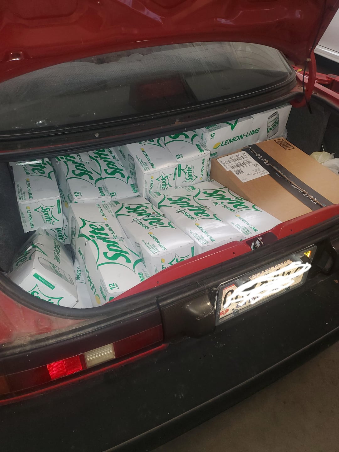 An open car trunk filled with multiple cases of Sprite soda cans and a cardboard box, with the license plate partially obscured.