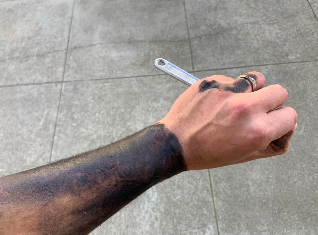 A person’s right hand and forearm are covered in black grease while holding a wrench over a concrete surface. The person is wearing two rings on their ring finger.