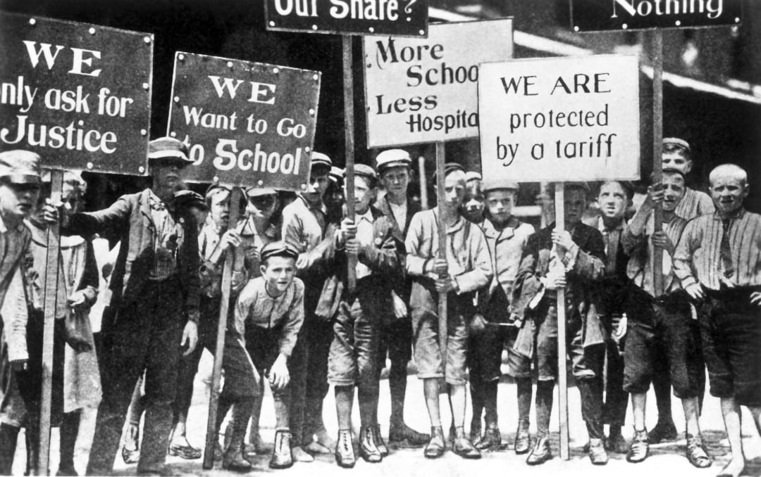 A group of boys in early 20th-century clothing hold protest signs with messages like "We want to go to school," "We only ask for justice," and "More school less hospital.