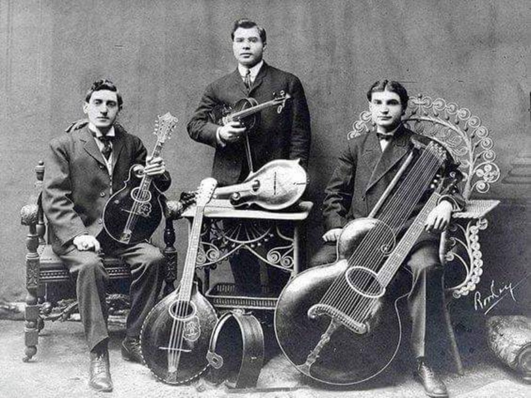 Three men in old-fashioned suits pose with unusual stringed instruments in a vintage black-and-white photograph. The men are seated and standing, surrounded by mandolins, guitars, and other unique string instruments.