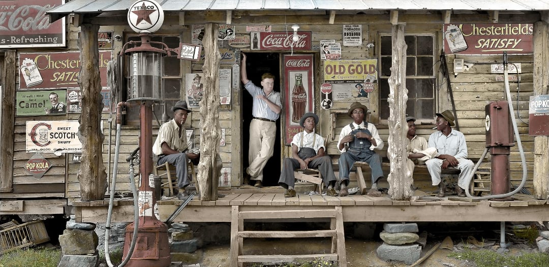 Five men sit and stand on the porch of an old wooden general store covered in vintage ads and signs, with a gas pump and weathered details visible in front. The scene appears to be from a past era.