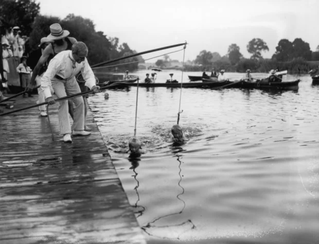 Two women participate in a 1920s swimming race with their heads in glass spheres attached to a frame, while a man monitors them from a dock; people watch from boats and the shore in the background.