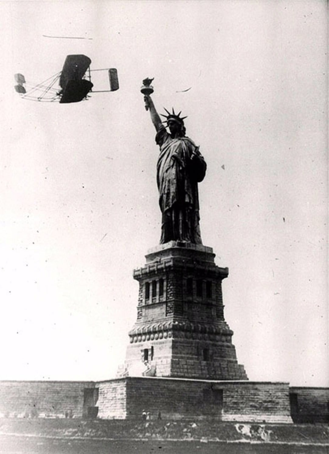 A vintage black-and-white photo shows an early biplane flying near the torch of the Statue of Liberty, which stands on its stone pedestal against a plain sky.