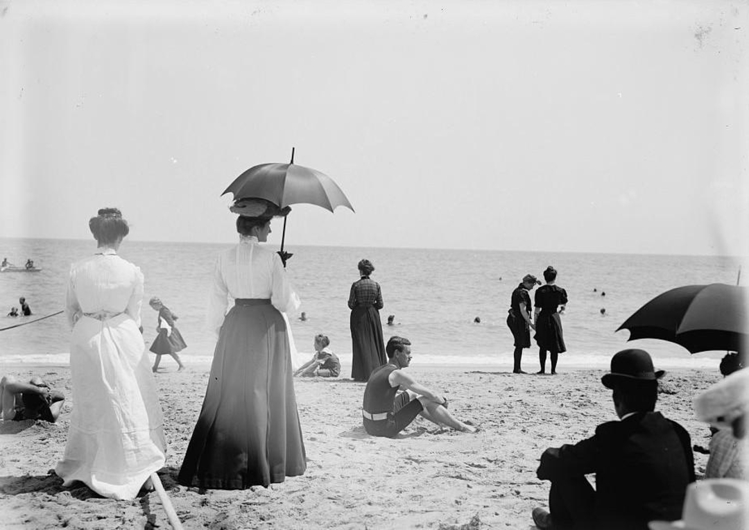 People in early 1900s clothing stand and sit on a beach. Women wear long skirts and hats, some holding parasols. Men wear suits and bowler hats. Some people are in the water, and the scene looks peaceful and relaxed.