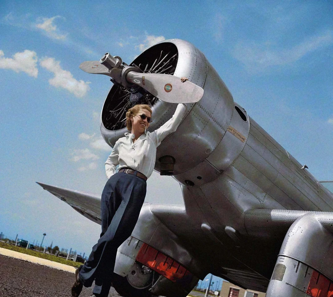 A woman in sunglasses leans against the propeller of a silver airplane on a sunny day, with a blue sky and a few clouds in the background. She wears a white shirt and dark trousers, standing confidently.