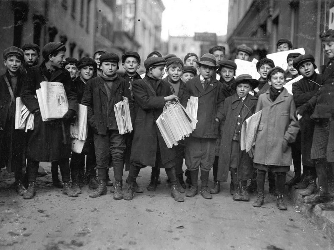 A group of boys, most holding newspapers, stand together on a city street in old-fashioned clothing and hats, suggesting they are newsboys from the early 20th century. The background shows tall buildings.