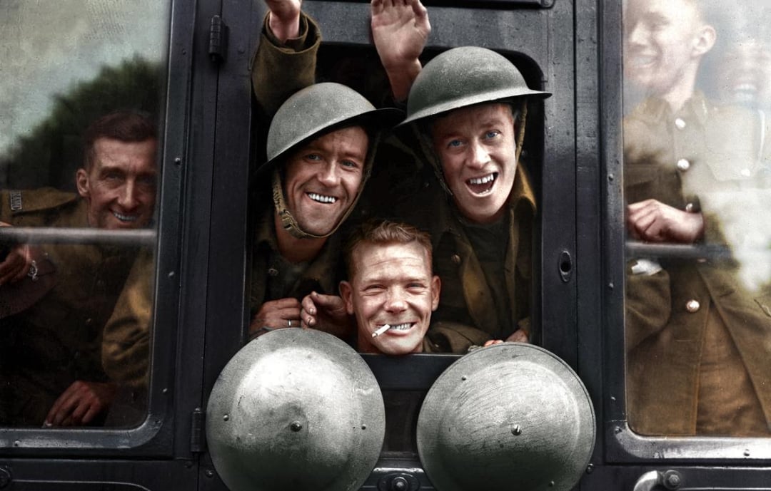 A group of smiling soldiers in uniform and helmets lean out of a train window, waving and looking cheerful as they prepare to depart.