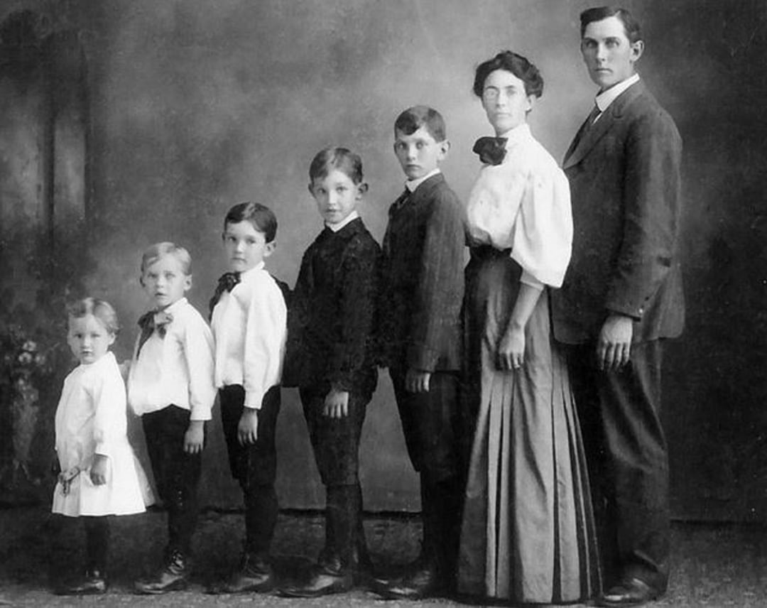 A vintage black-and-white photo of a family of six, lined up from youngest to oldest. Three boys, one girl, and two adults stand in profile, dressed in early 20th-century clothing, against a plain studio backdrop.