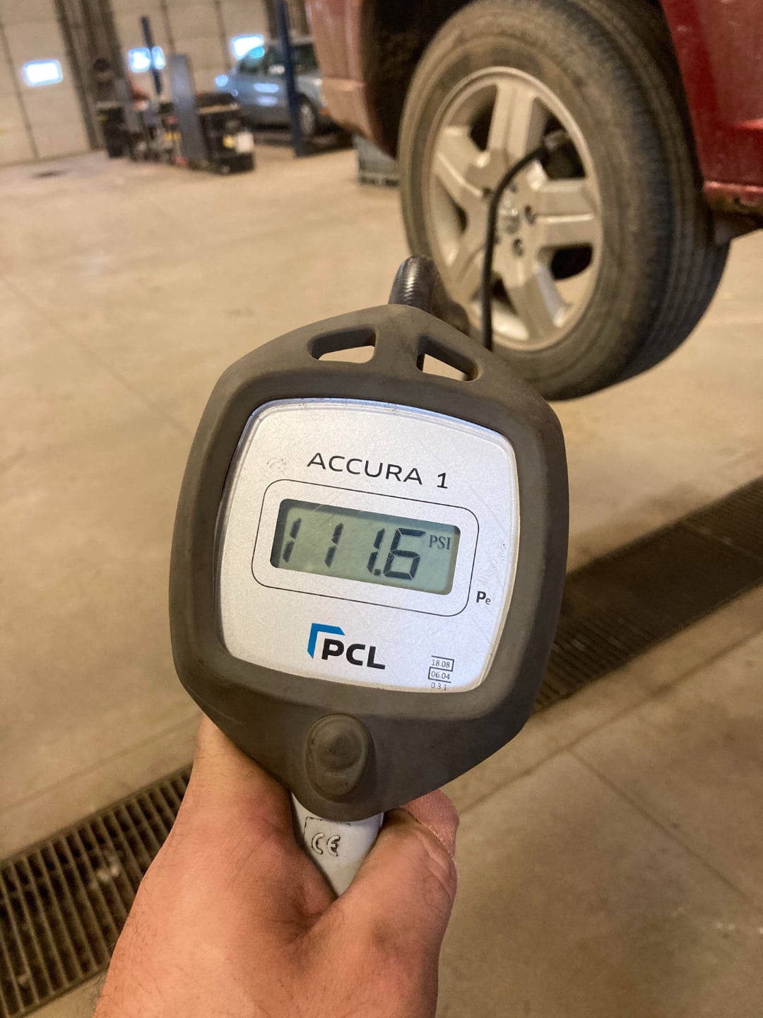 A hand holds a digital tire pressure gauge reading 11.6 PSI in front of a raised car wheel inside an auto repair shop.