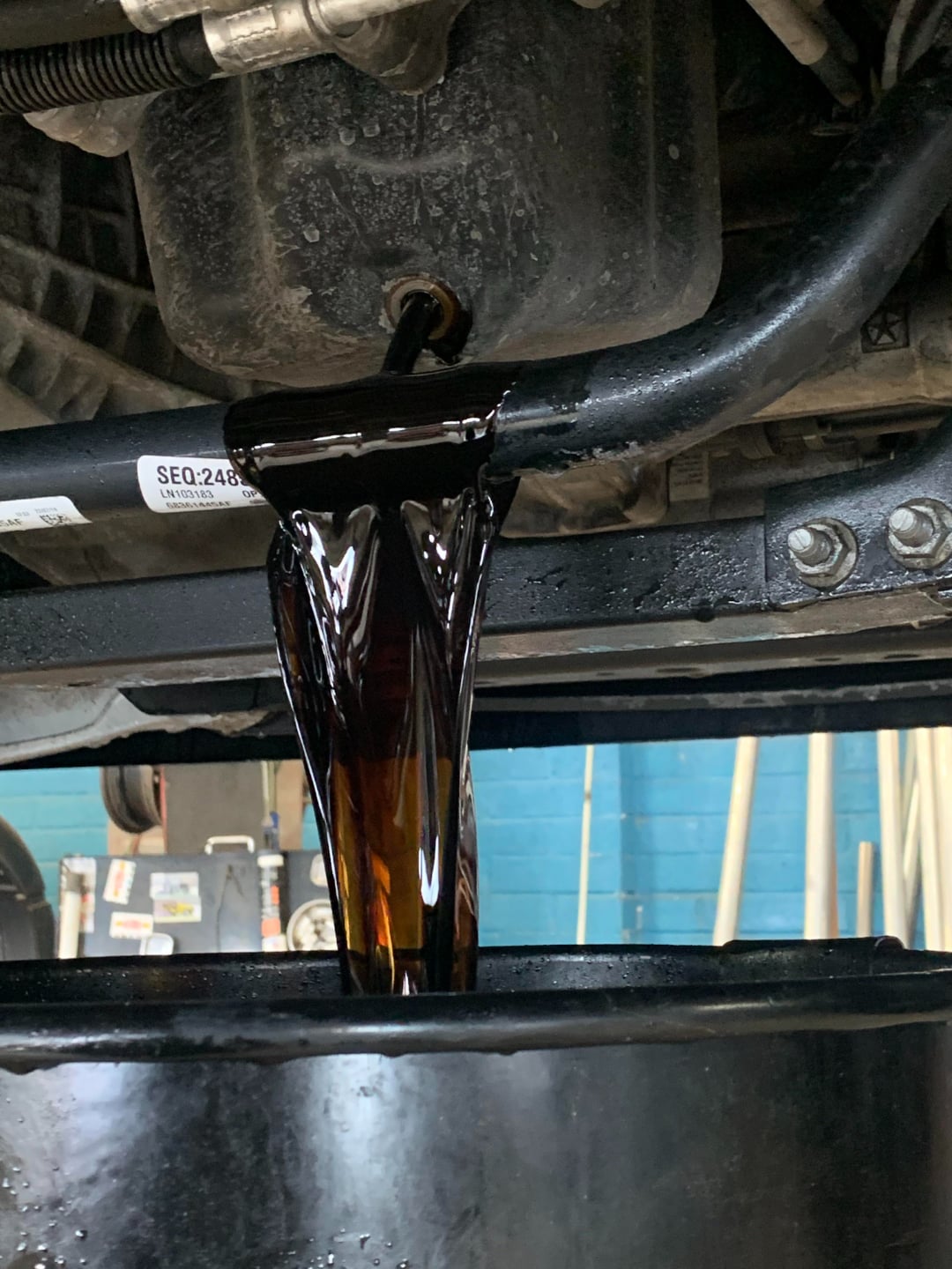 Thick, dark engine oil is flowing from a car's oil pan drain plug into a black container during an oil change in a workshop with a blue wall in the background.