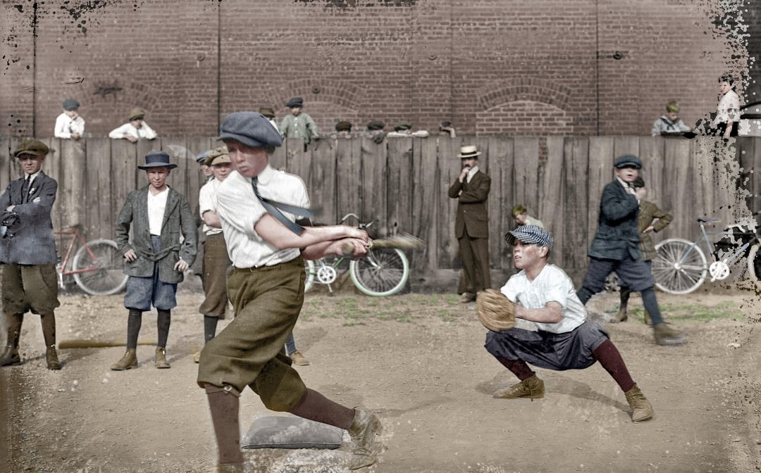 A group of boys dressed in early 20th-century clothing play baseball on a dirt lot, with a batter swinging at a pitch and other boys watching or playing in the background. Bicycles and a brick wall are visible behind them.