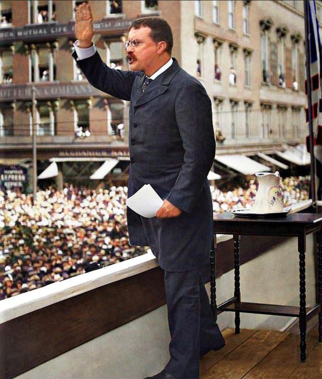 A man in a dark suit stands on a balcony, raising his right hand as he addresses a large crowd gathered below. He holds papers in his left hand and stands beside a small table with a pitcher and glass.