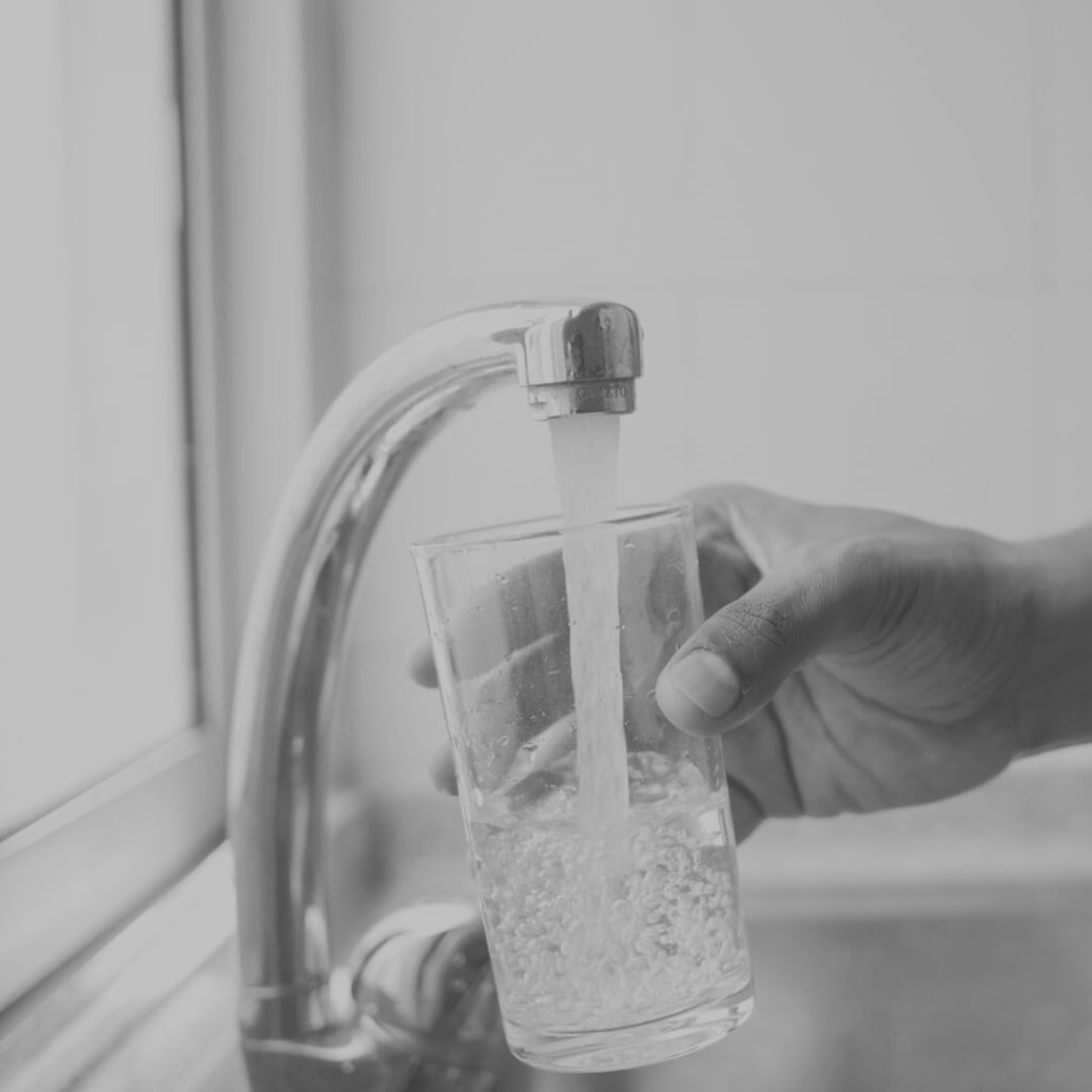 A hand is holding a clear glass under a running kitchen faucet, filling it with water. The scene takes place in front of a window, and the image is in black and white.