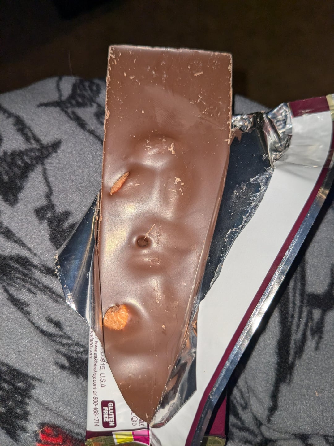 A close-up of a partially unwrapped chocolate bar with whole nuts visible inside, resting on a gray fabric with a black animal pattern.