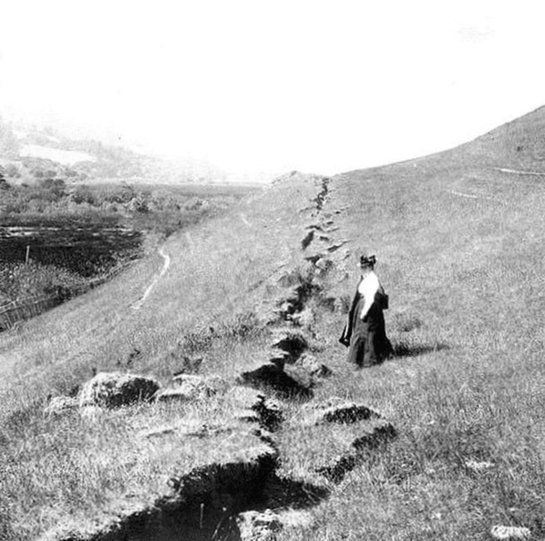 A woman in early 20th-century clothing stands beside a deep crack in grassy hills, marking the earthquake rupture of the San Andreas Fault in California. The landscape is uneven and sparsely vegetated.