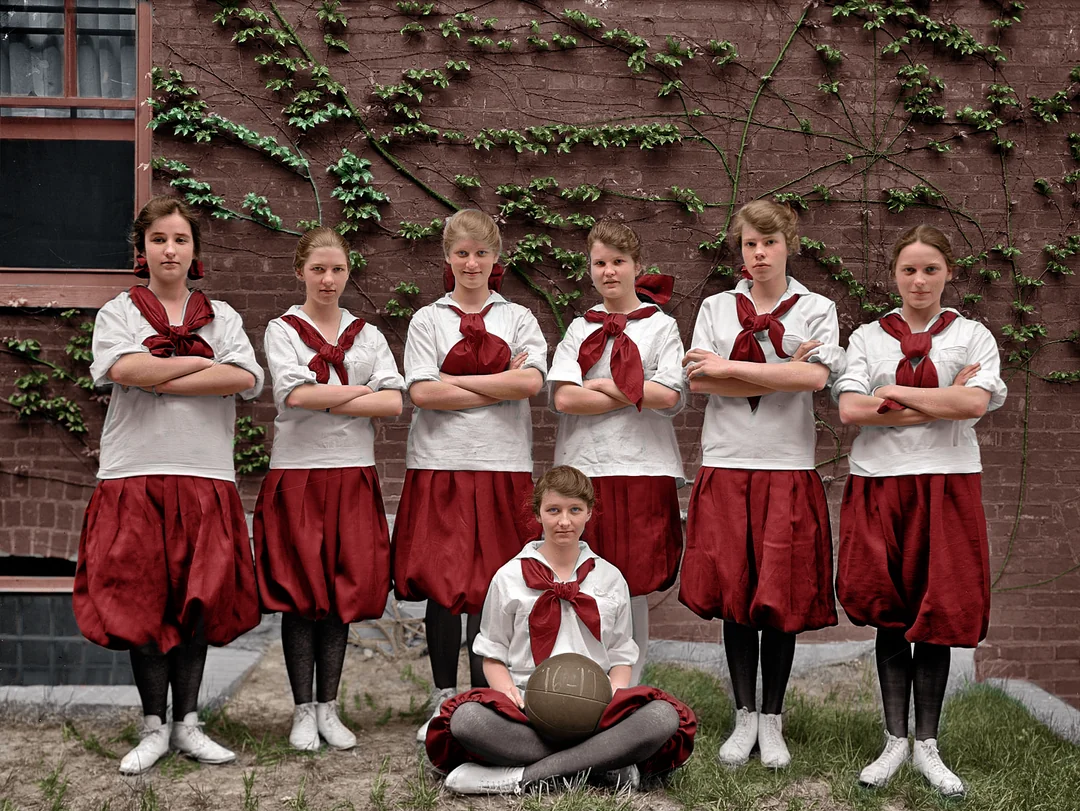Seven girls in vintage gym uniforms pose in front of a brick wall, six standing with arms crossed and one sitting cross-legged in front, holding a basketball. Green vines grow on the wall behind them.