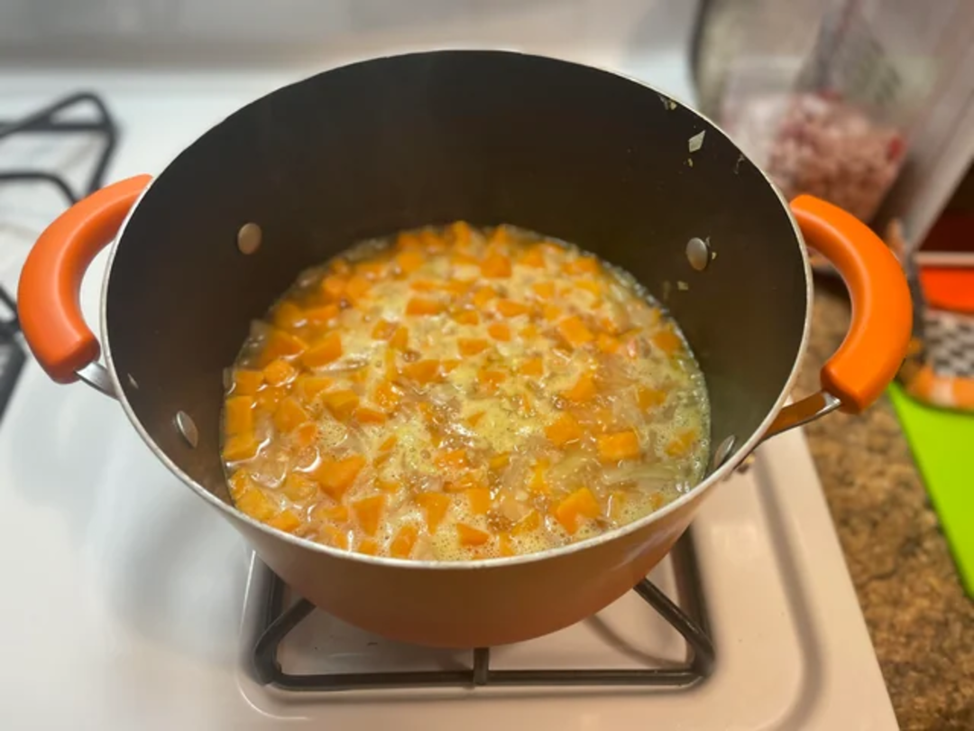 A large orange pot on a stove contains a simmering mixture of diced carrots and onions in a broth. The countertop nearby holds kitchen tools and chopped ingredients.