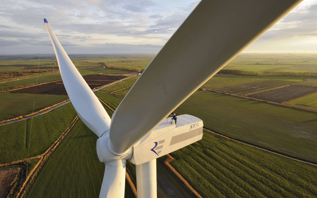 A person in a blue jacket stands on top of a large white wind turbine, overlooking green fields and farmland under a cloudy sky at sunset.