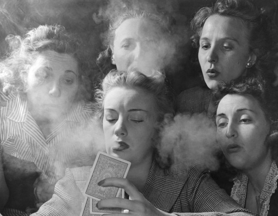 Five women, surrounded by swirling smoke, sit closely together as one woman holds playing cards. The group appears engaged and focused, with smoke partially obscuring their faces.