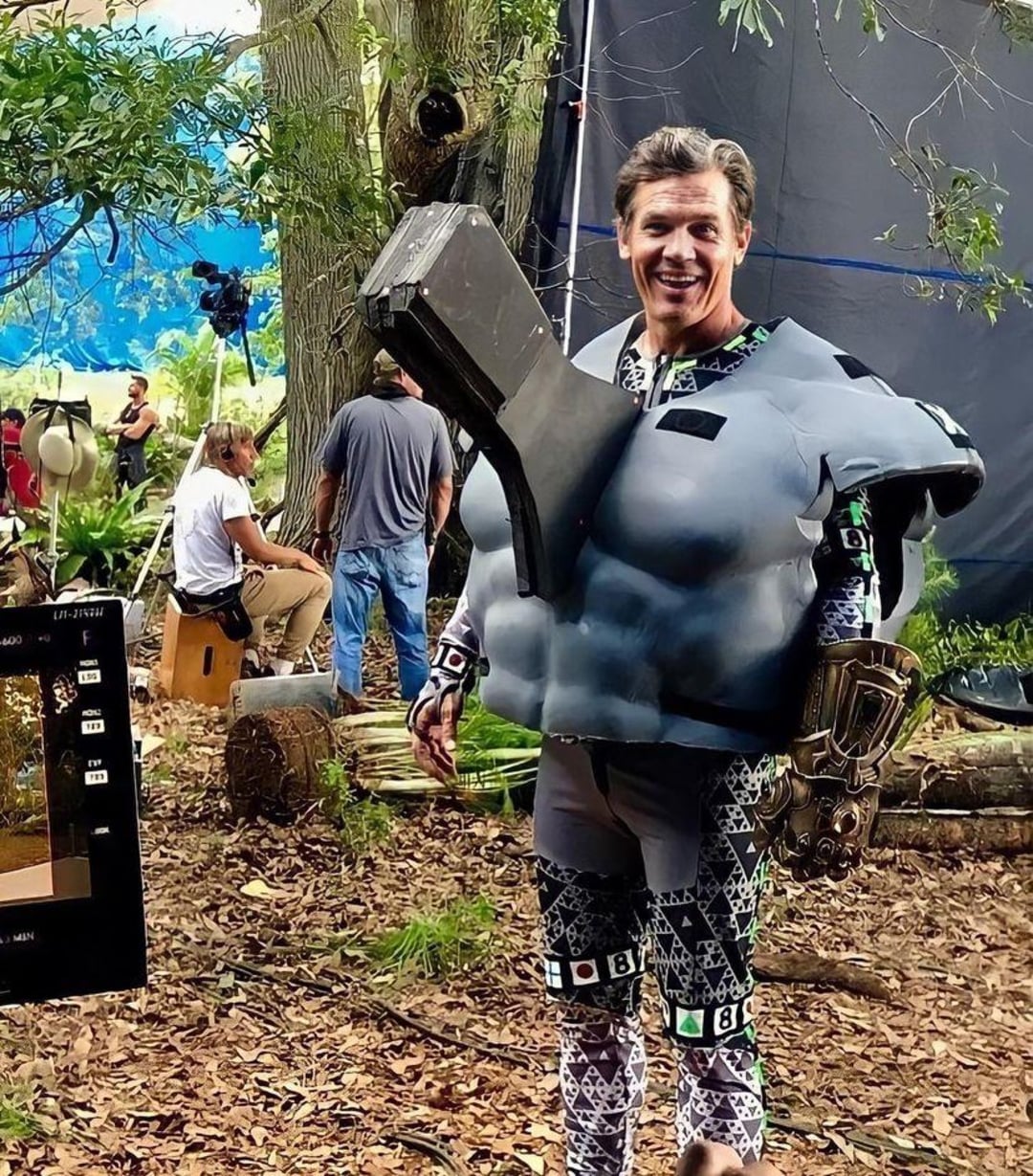 A smiling man wearing a bulky, gray motion-capture suit with a large foam torso and an arm extension stands in a wooded film set, surrounded by crew members and equipment.