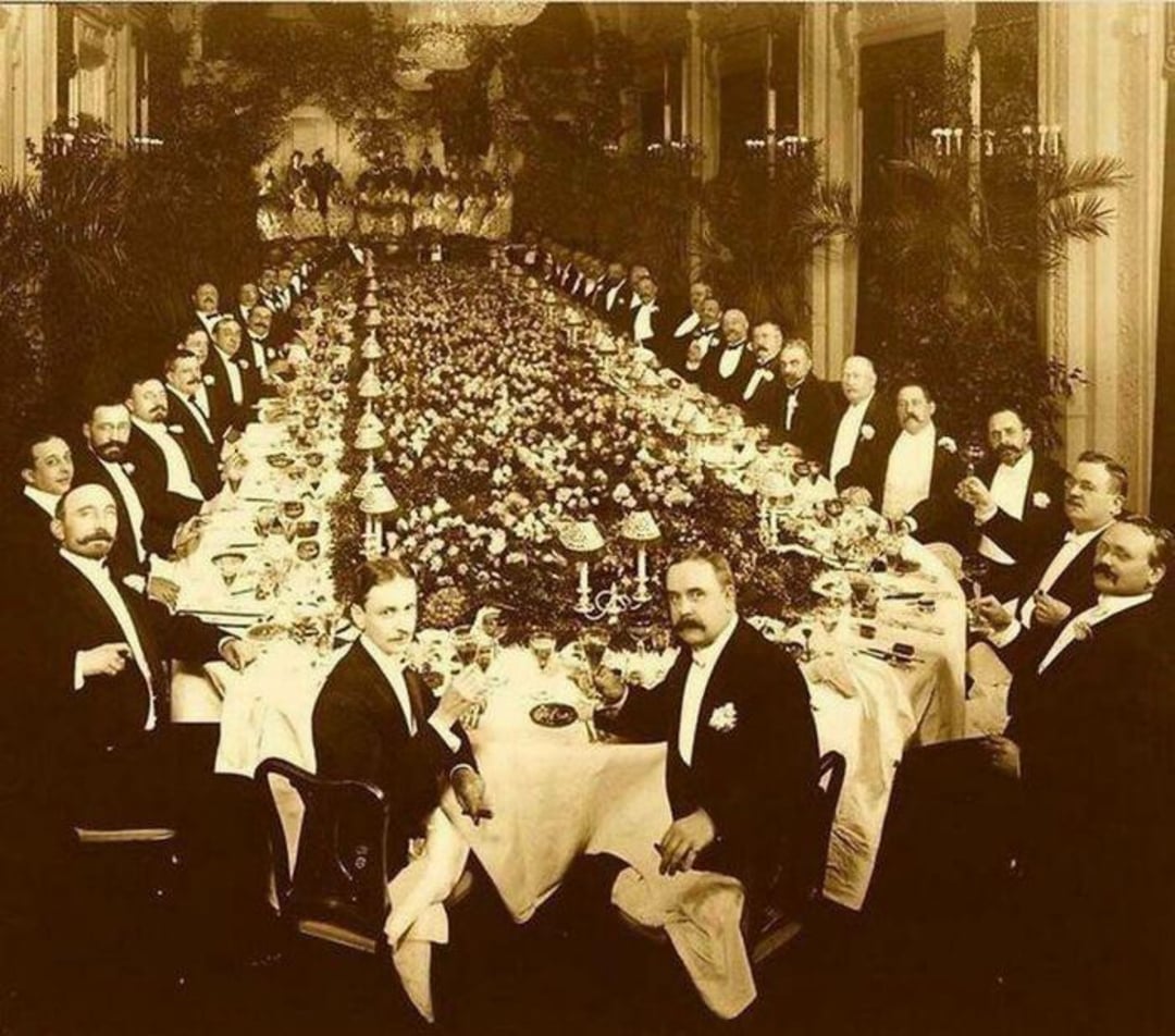 A sepia-toned photo of a large formal banquet with many men in tuxedos seated around a long, flower-adorned table in an ornate room, raising glasses and facing the camera.