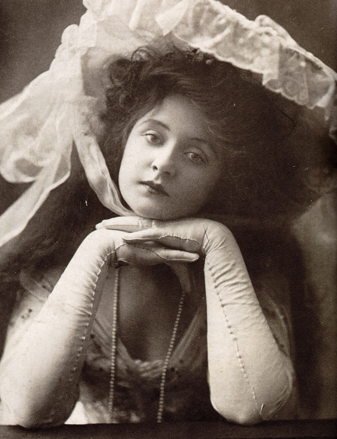 A young woman in vintage attire poses with her chin resting on her gloved hands, wearing a large, decorative hat with ribbons and lace; the image is in black and white with a soft, elegant feel.