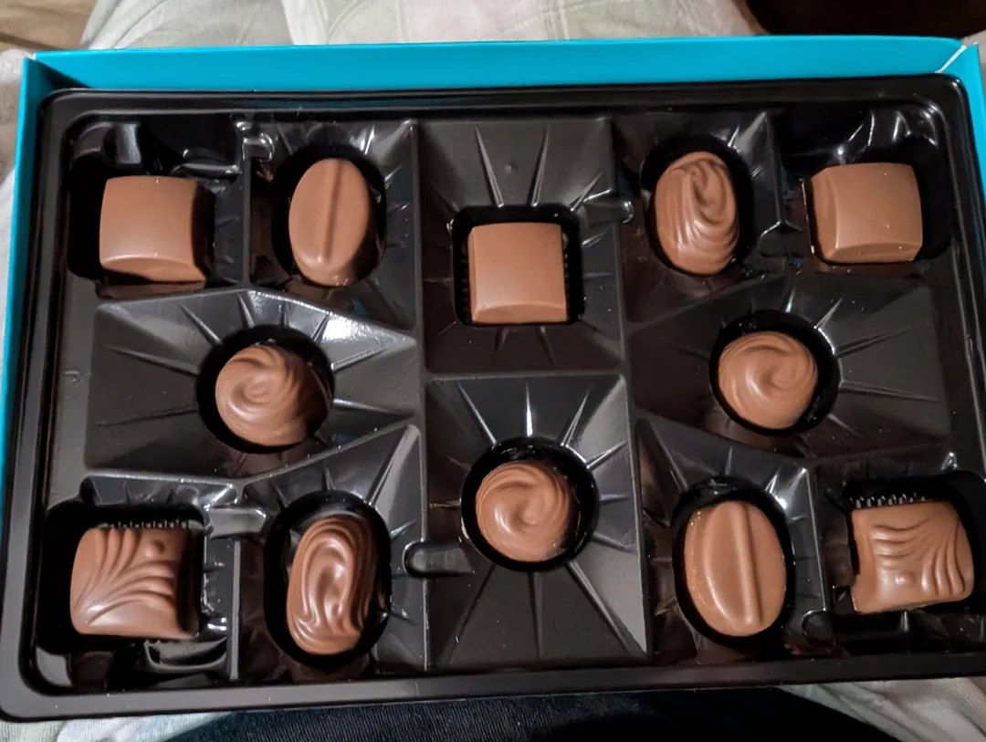 A partially empty box of assorted milk chocolates, with several chocolates missing and only seven pieces remaining in a black plastic tray inside a blue box.